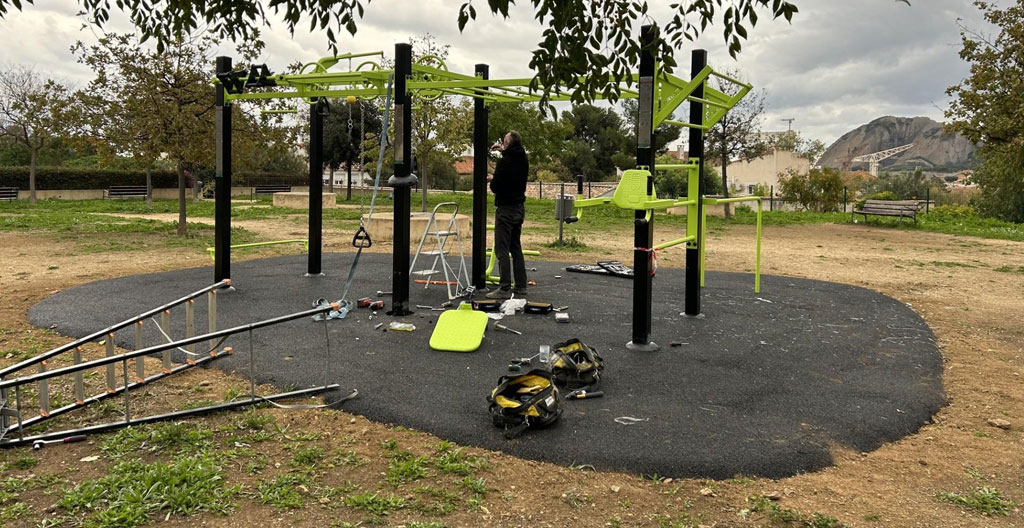 Technicien installateur en vêtements de travail fixant la base d'un agrès de fitness extérieur sur dalle béton, outils professionnels à proximité, chantier d'aménagement sportif en arrière-plan